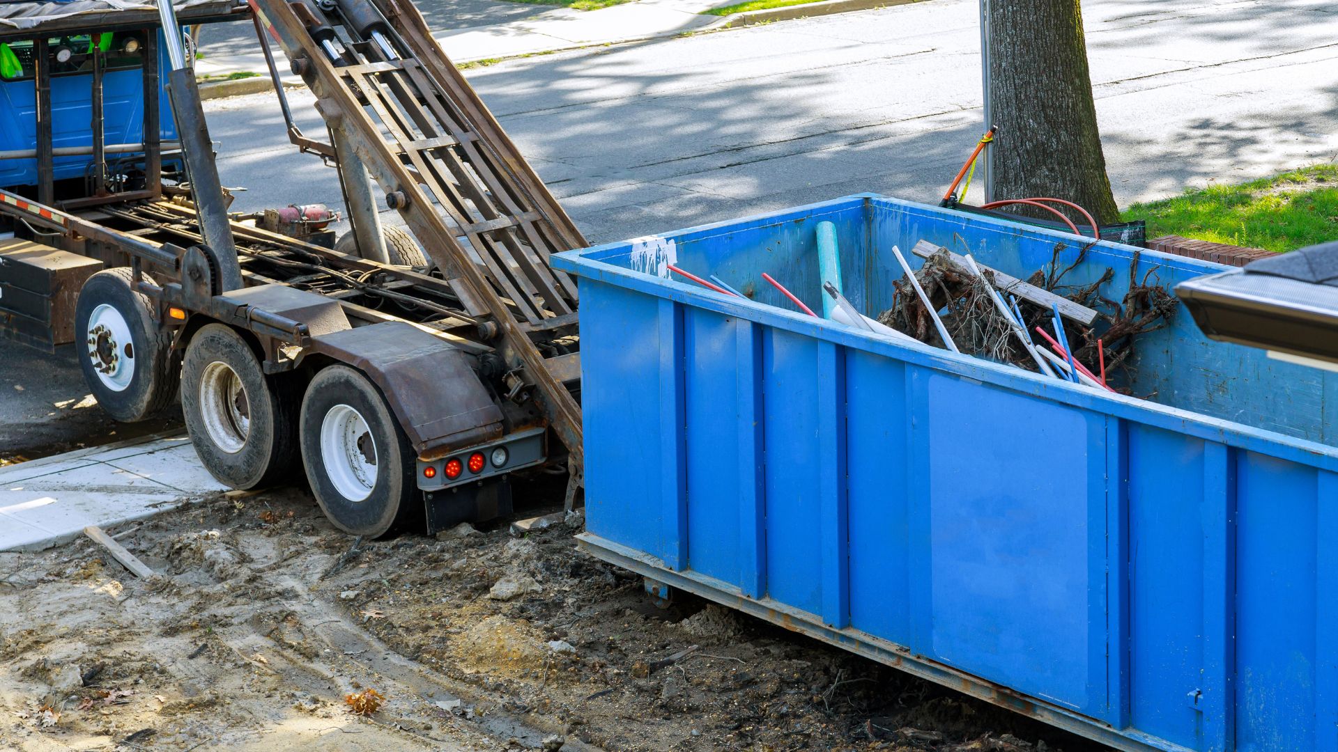 Blue dumpster and truck parked on dirt near street with debris